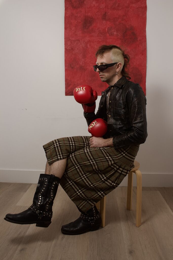 eli sitzt vor einem roten gemälde auf einem stuhl udn trägt stiefel sowie rote boxhandschuhe