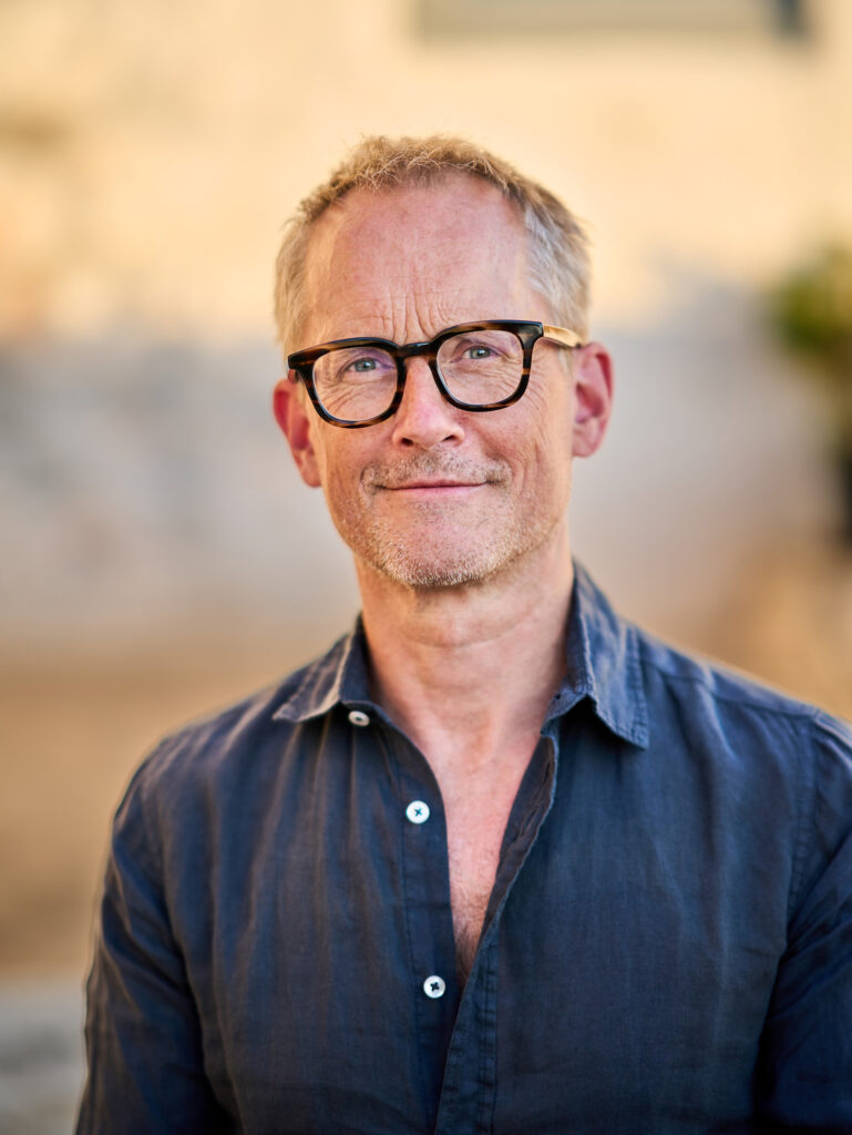 text. ein mann mit dunklem hemd, dreitagebart und makanter, dunkler brille im portrait. er lächelt leicht und der hintergrund ist verschwommen.