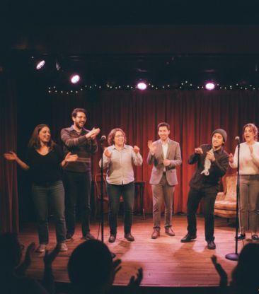 sechs menschen auf einer bühe mit roten samtvorhängen, die mit mikrofonen theater spielen und dabei spass haben